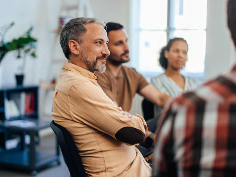 Behavioral health patient smiling during group therapy session in bright, modern facility with other participants in background Behavioral health patient smiling during group therapy session in bright, modern facility with other participants in background