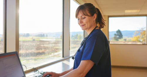Female nurse in blue scrubs with stethoscope working at a computer workstation in a rural hospital facility