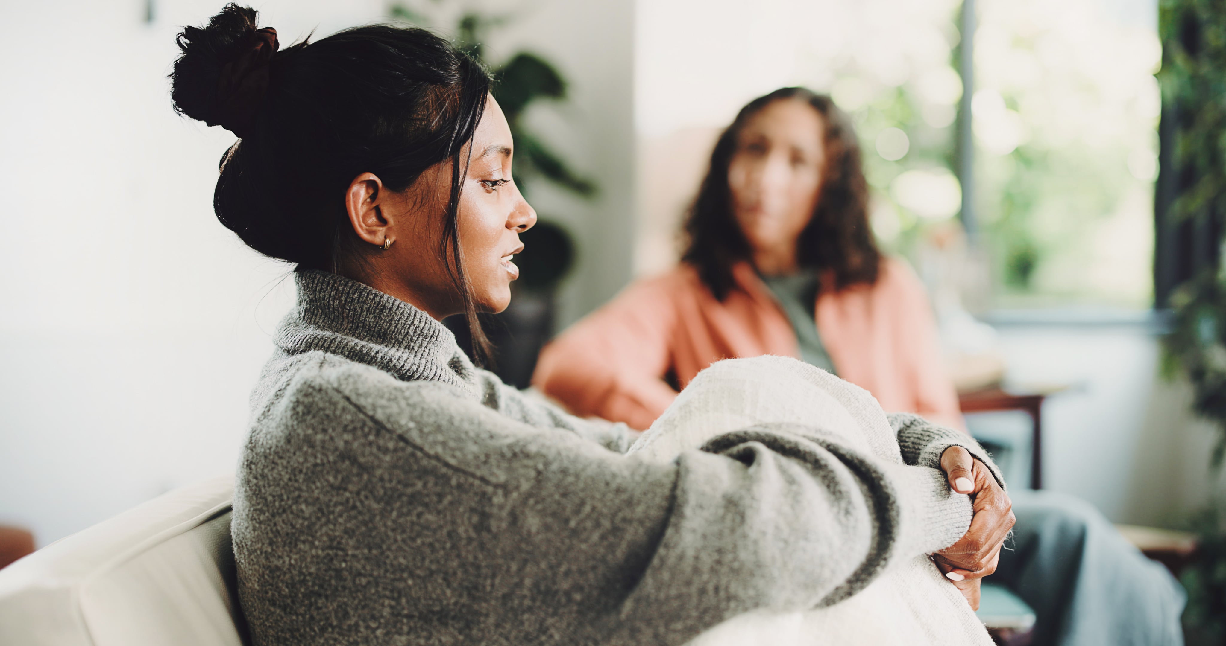 A woman in a gray sweater sits thoughtfully during a one-on-one therapy session, with a counselor listening attentively in the background