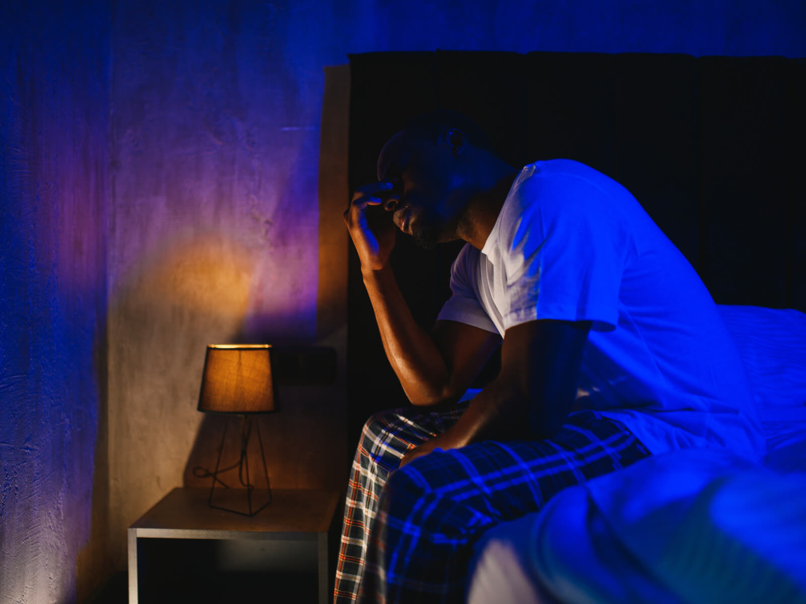 Person with fatigue and insomnia, sitting on bed in dimly lit room with blue lighting, head in hands, appearing distressed next to small bedside lamp.