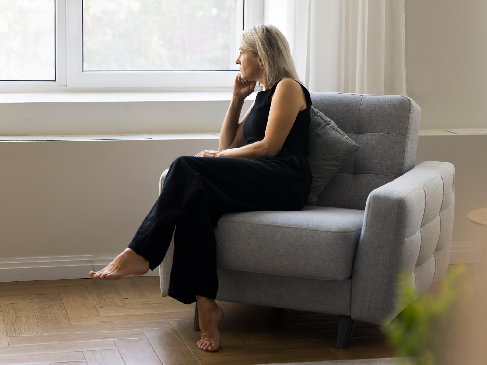 CEO stressed from work burnout in black outfit sitting in gray armchair, gazing thoughtfully out of window in bright minimalist room with wooden floor.